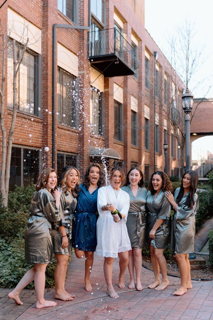 Bride & bridesmaid pop the champagne in the garden courtyard at their industrial chic warehouse style wedding venue near Winston Salem 