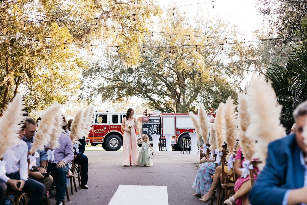 Bridesmaid dressed in pink & flower girl dressed in green walk down the aisle of a garden wedding ceremony at Rustic Grace Barn near Orlando